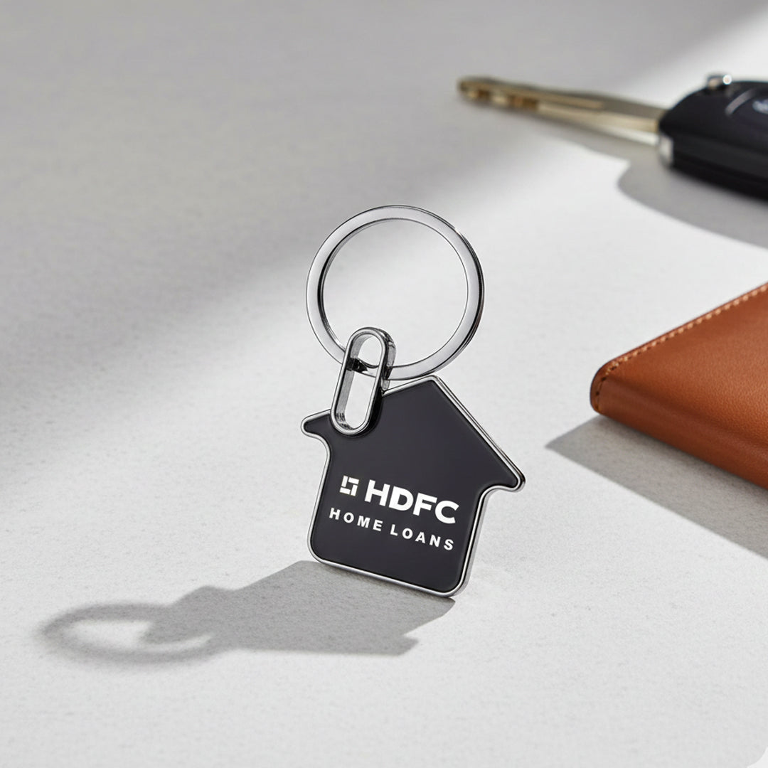 The Home Metal Key Chain - AIKC - 2505, shaped like a house and printed with HDFC Home Loans, stands upright beside a brown wallet and car keys—ideal as a realtor client gift.