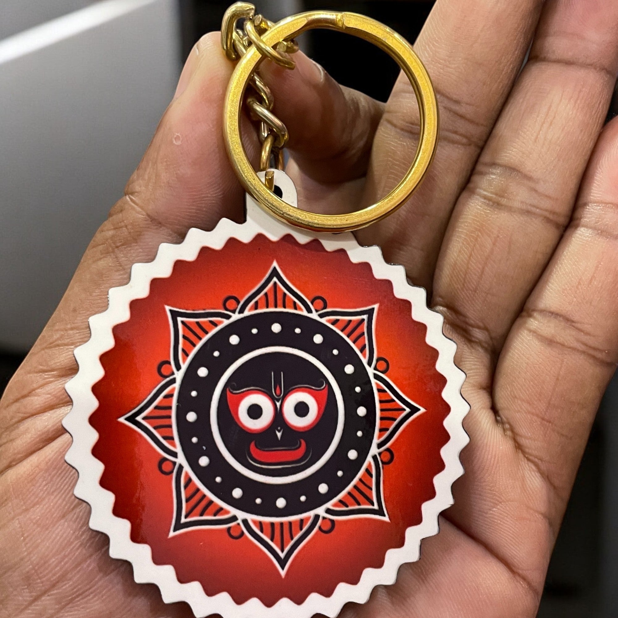 A hand holds a Jai Jagannath Key Chain (Set of 3) with a circular design showing a stylized black and white face with red eyes on a red background, accented by a decorative border.