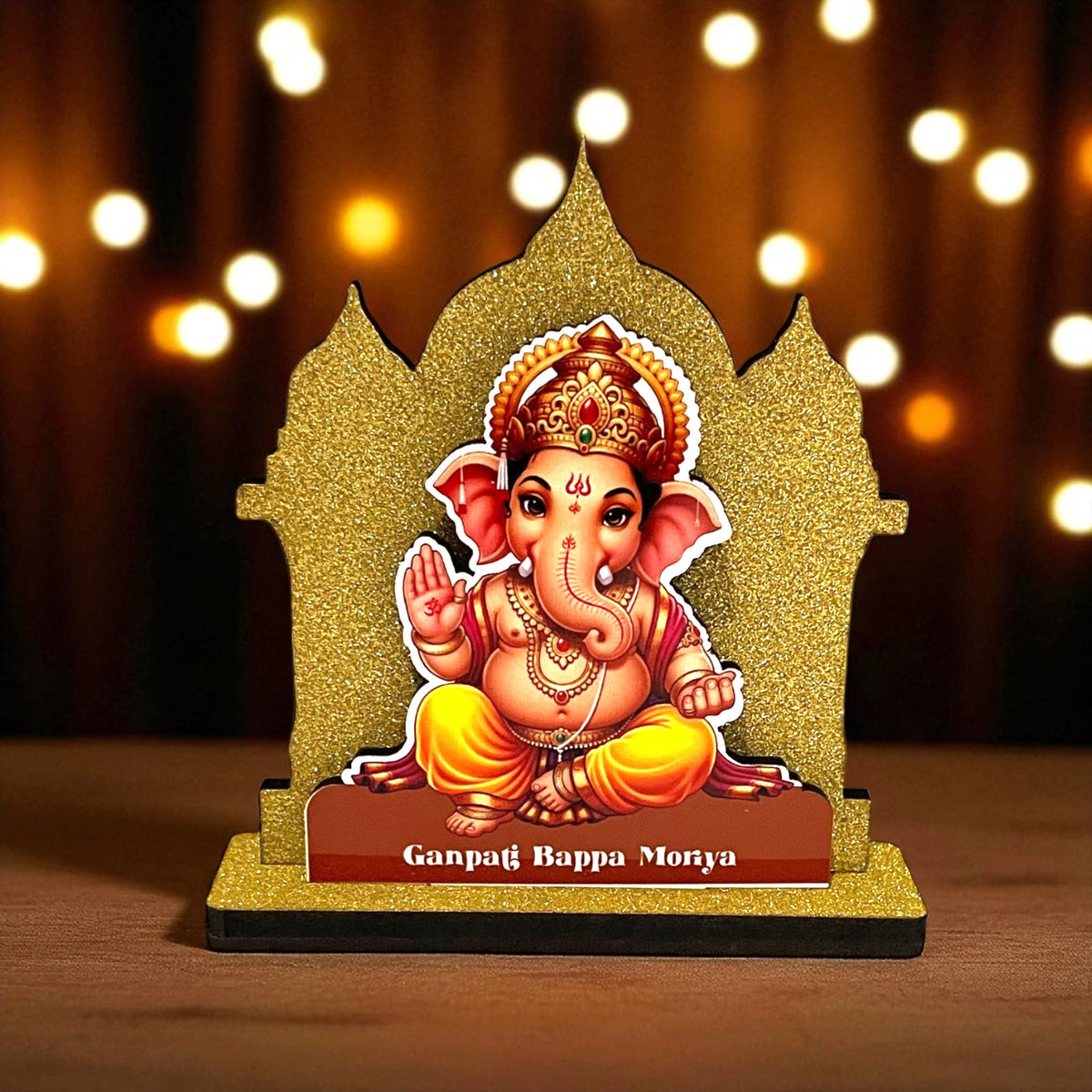 The Hindu Gods Mini Temple features a decorative idol of Lord Ganesha on a stand inscribed with "Ganpati Bappa Morya," set against a glittery gold backdrop, perfect for your car dashboard. Warm, blurred lights glow in the background.